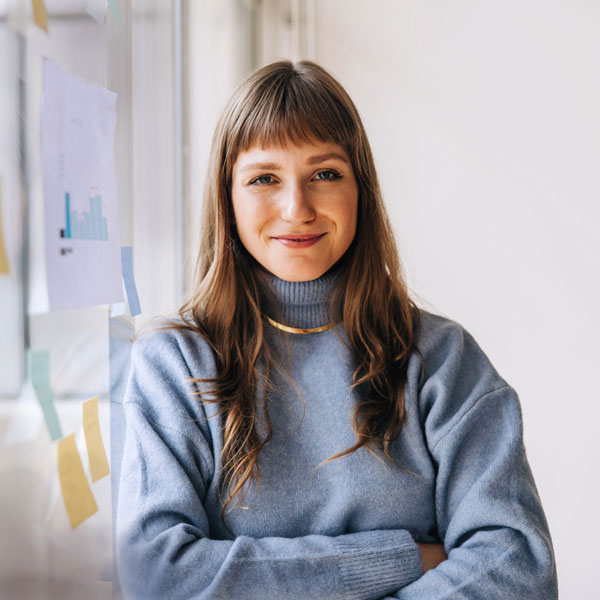 Junge Frau im Büro Junge Frau mit langen braunen Haaren und blauem Pullover steht lächelnd in einem hellen Büro mit Notizzetteln und Diagrammen an der Wand – Symbol für mentale Gesundheit und Wohlbefinden am Arbeitsplatz.